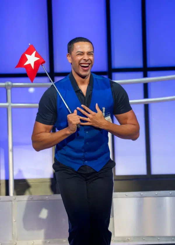 A smiling person wearing a blue vest holds a small red flag with a white star. The background features blue-lit panels and metal railing, suggesting a stage or performance setting.