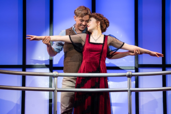 A man stands behind a woman on a ships deck, holding her outstretched arms. They smile at each other, recreating the iconic scene from Titanic. The background is lit blue with window panels.