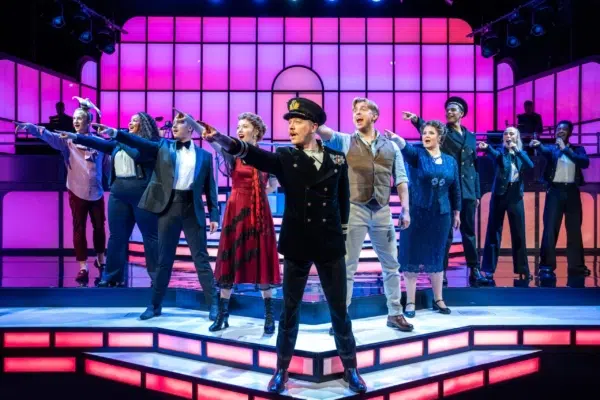 A group of performers in colorful, vintage-inspired costumes stand on a lit stage with bright pink and purple lights, dramatically pointing forward with confident expressions during a musical theater performance.