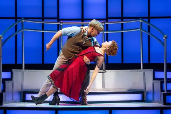 A male actor dips a female actor in a dramatic dance pose on stage, with a modern blue and purple-lit backdrop and a metallic platform behind them. The woman wears a red dress, and the man wears a vest and light shirt.