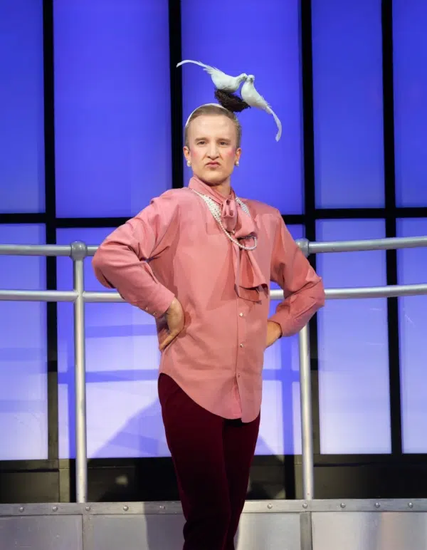 A person wearing a pink blouse and burgundy pants stands with hands on hips in front of a blue-lit background. A white dove is perched on their head, and they have a playful expression.